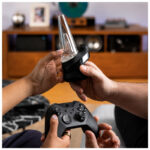 Top-down lifestyle photo of two people gaming in a cosy living room while passing a black Puffco Peak vaporizer between them, with a game controller in the foreground and warm natural lighting.