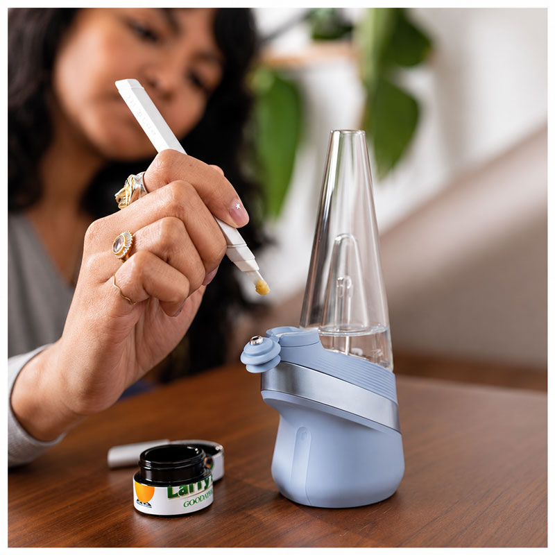 Close-up lifestyle photo of a person loading concentrate into a blue Puffco Peak vaporizer with a dab tool on a wooden table indoors.
