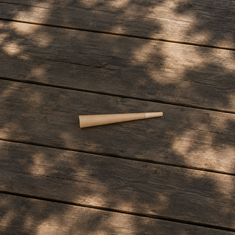 Top-down outdoor photo of a single pre-rolled cone resting on a weathered wooden table in natural sunlight with soft tree shadows.