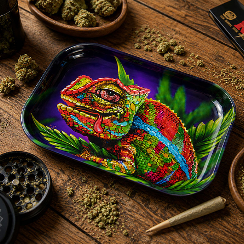 Top-down view of a colourful psychedelic chameleon rolling tray on a wooden table, surrounded by ground herbs, cannabis buds, a grinder, rolling papers and a rolled joint.