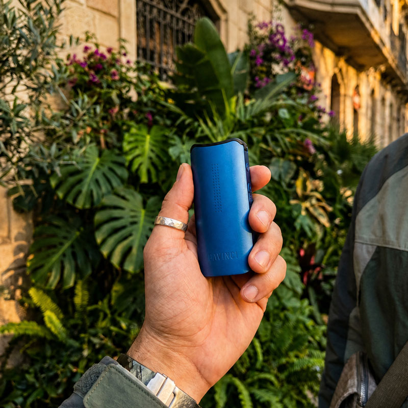 Blue DaVinci IQ Core vaporizer held in a hand outdoors against a leafy background