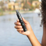 Black XMax V4 Pro dry herb vaporizer held in one hand beside a lake